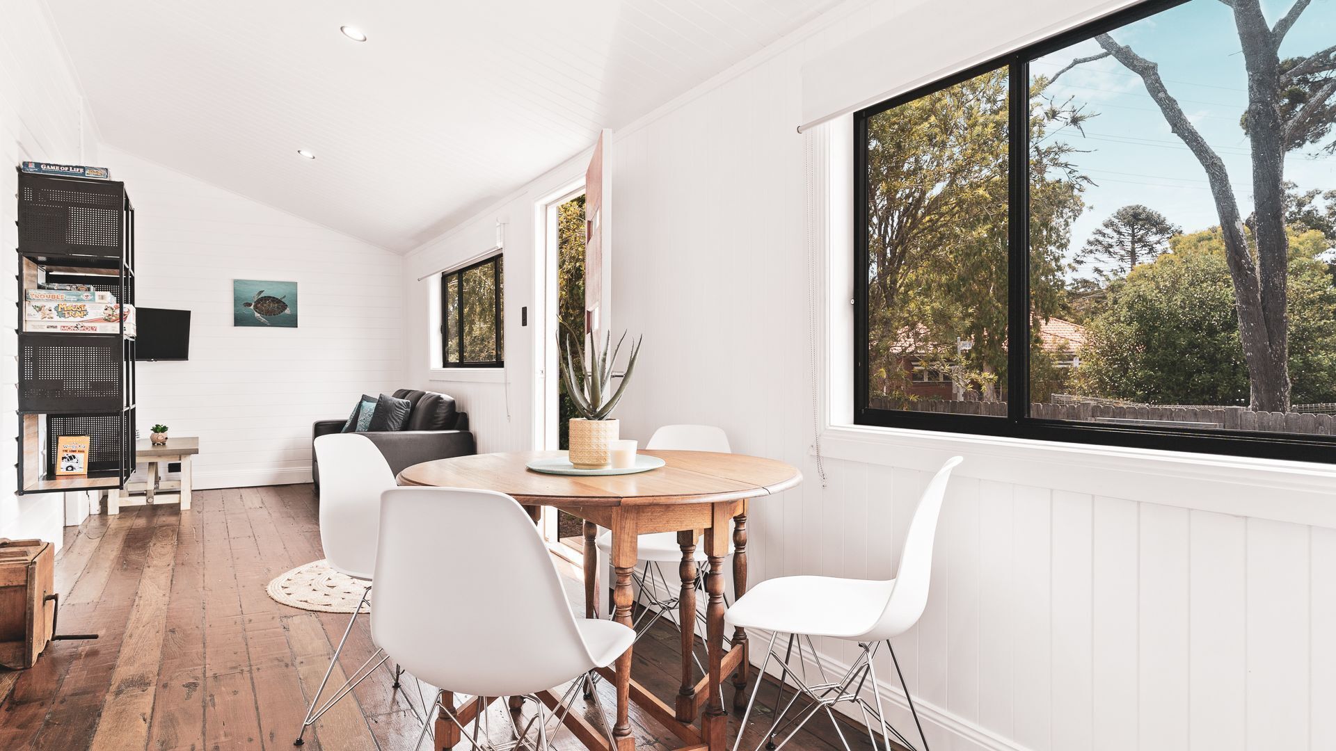 A modern dining area with a wooden table and white chairs, a bookshelf, and a view of trees through a large window.