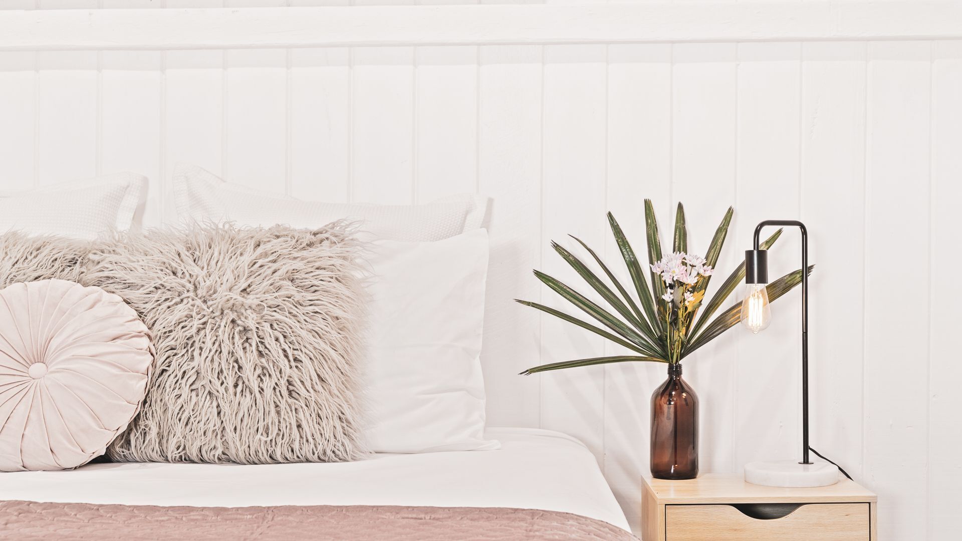 A neatly made bed with fluffy pillows, a bedside table with a vase of flowers and a lamp, and a white wall with vertical paneling.