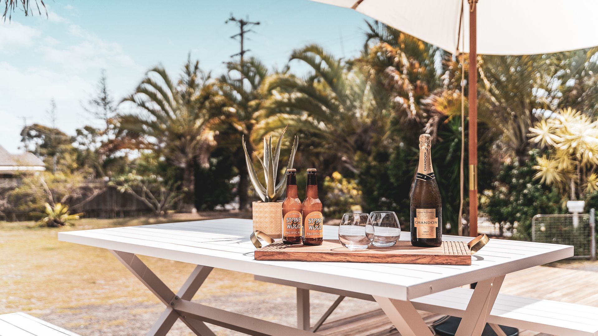 An outdoor table with a bottle of Chandon wine, two Stone Wood Pacific Ale bottles, and glasses.