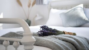 Un lit avec une couverture grise, une tête de lit blanche et un bouquet de fleurs de lavande.