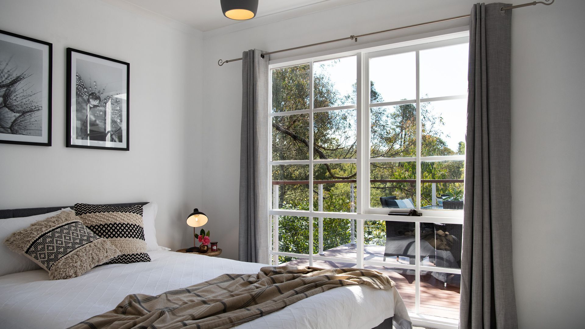Een slaapkamer met een groot raam, een bed met gestreepte kussens, een bedlampje en twee ingelijste foto's aan de muur.