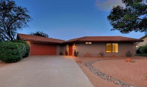 A two-story house with a red-tiled roof, a driveway, and a landscaped front yard with rocks and shrubs.