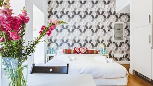 Habitació d'hotel moderna amb paret de disseny geomètric, llit amb roba blanca, tauleta de nit amb làmpada i gerra amb flors roses.