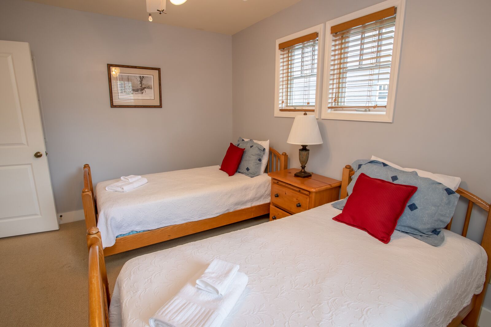 A bedroom with two single beds, a nightstand with a lamp, a framed picture on the wall, and a window with blinds.