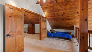 Quarto com teto inclinado, paredes de madeira, cama com roupa de cama azul e ventoinha no teto.