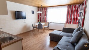 A modern living room with a grey sofa, a dining table with chairs, a flat-screen TV mounted on the wall, and a window with red and white curtains. - Ideal for mountain lovers, couples and small families  (6653 Bach)