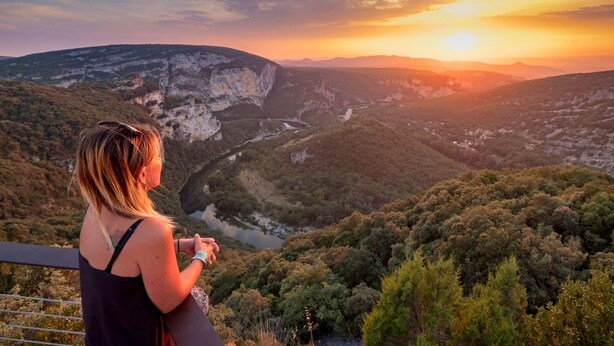 Eine Person blickt auf ein malerisches Tal mit Fluss und Klippen bei Sonnenuntergang.