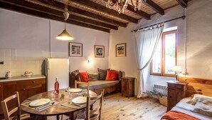Ein gemütliches Zimmer mit einem Holzesstisch, gedeckt für zwei Personen, einer Couch mit Kissen, einem kleinen Schrank und einem Fenster mit Vorhängen.