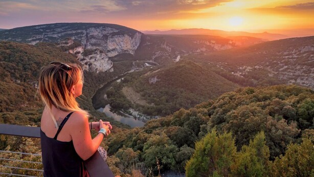 Eine Person blickt über ein Tal mit Fluss und steilen Felswänden bei Sonnenuntergang.
