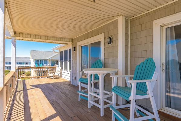 A covered outdoor area with a wooden deck, two white tables, and two turquoise Adirondack chairs. - Over The Moon - 4 BR home, Oceanfront B-Lot with Amazing Views! Dog Friendly! (Surf City)