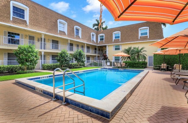A swimming pool with a metal ladder, surrounded by a brick patio. There are outdoor tables and chairs under orange umbrellas. - PIERRE CLUB 14 - Updated Tropical Condo, Best Location in Olde Naples! (Naples)