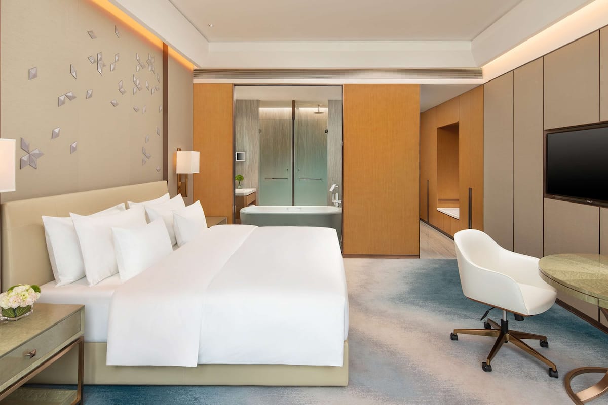 A modern hotel room with a large bed, a desk, a chair, a TV, and a bathroom visible through an open door.