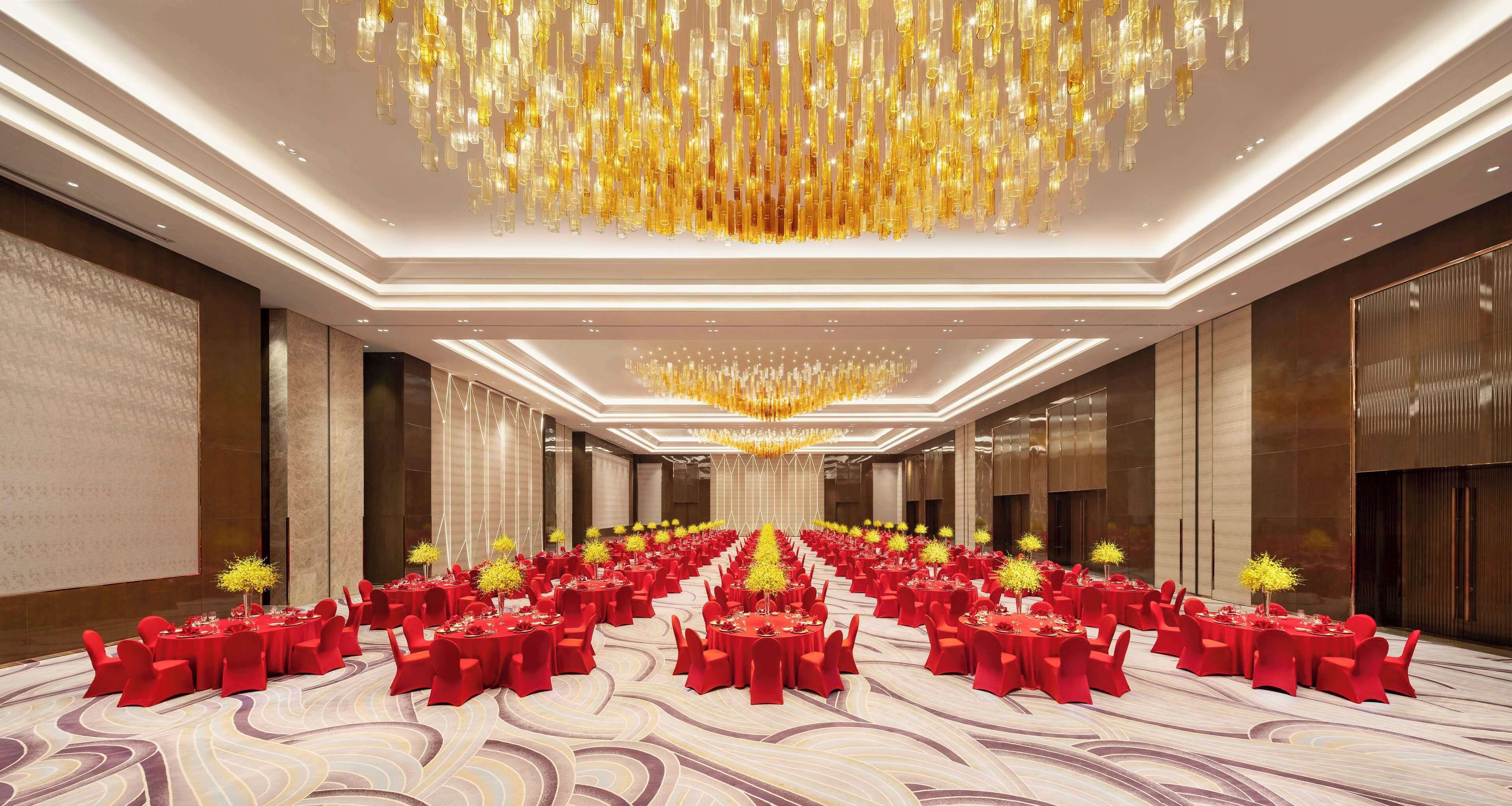 A spacious banquet hall with rows of round tables, red chairs, and a central chandelier.