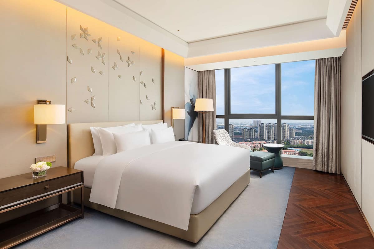 A modern hotel room with a large bed, two bedside lamps, a flat-screen TV, a city view, and a small side table.