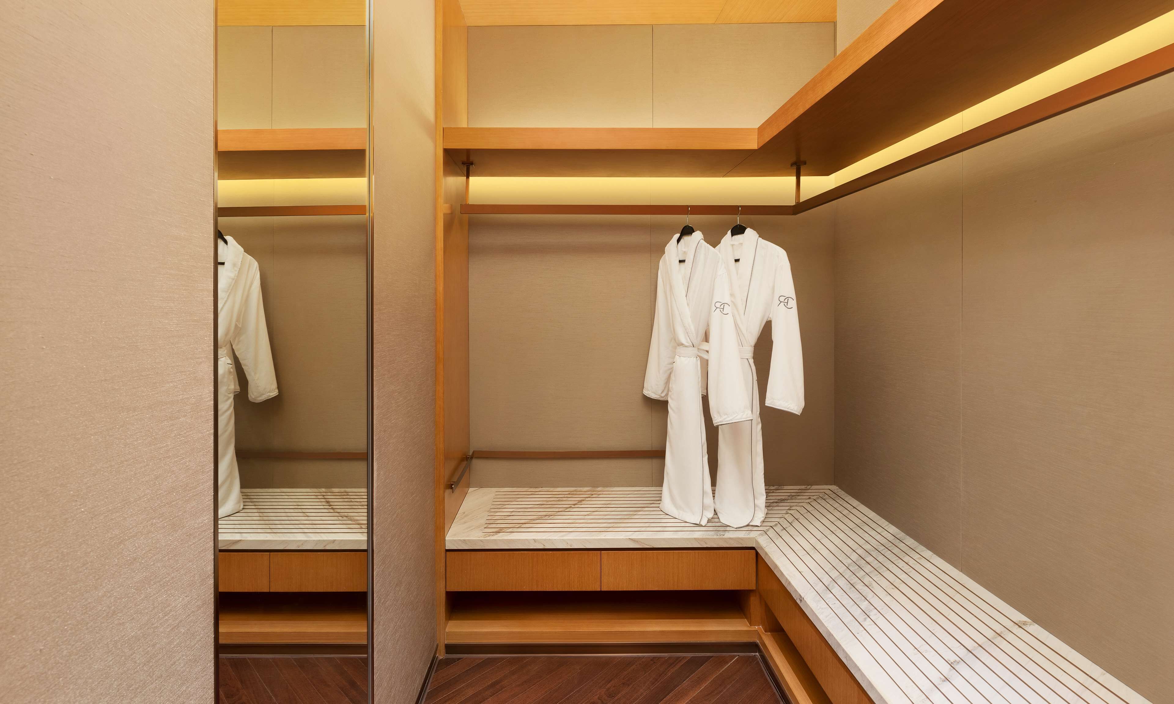 A modern, minimalist bathroom with wooden flooring, a bench, and two white robes hanging on hooks.