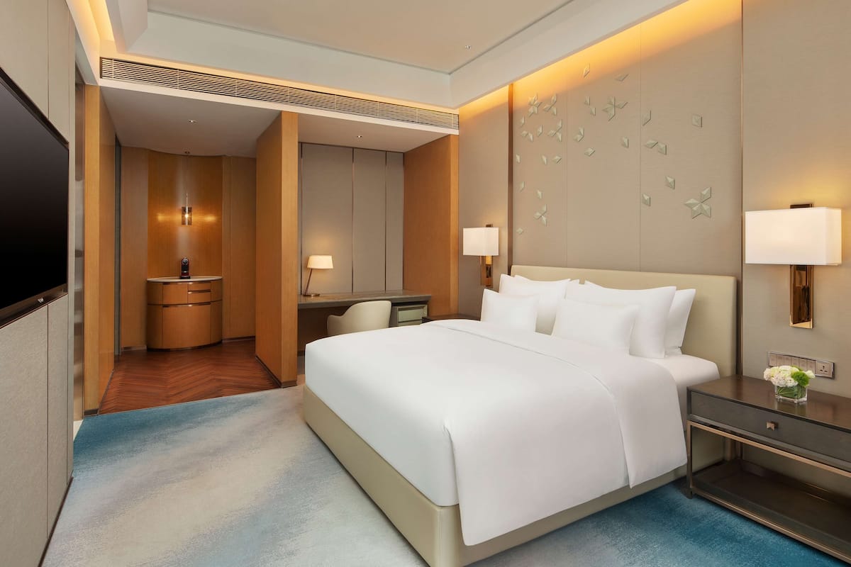 A hotel room with a large bed, a desk with a chair, a TV mounted on the wall, and a bedside table with a lamp.