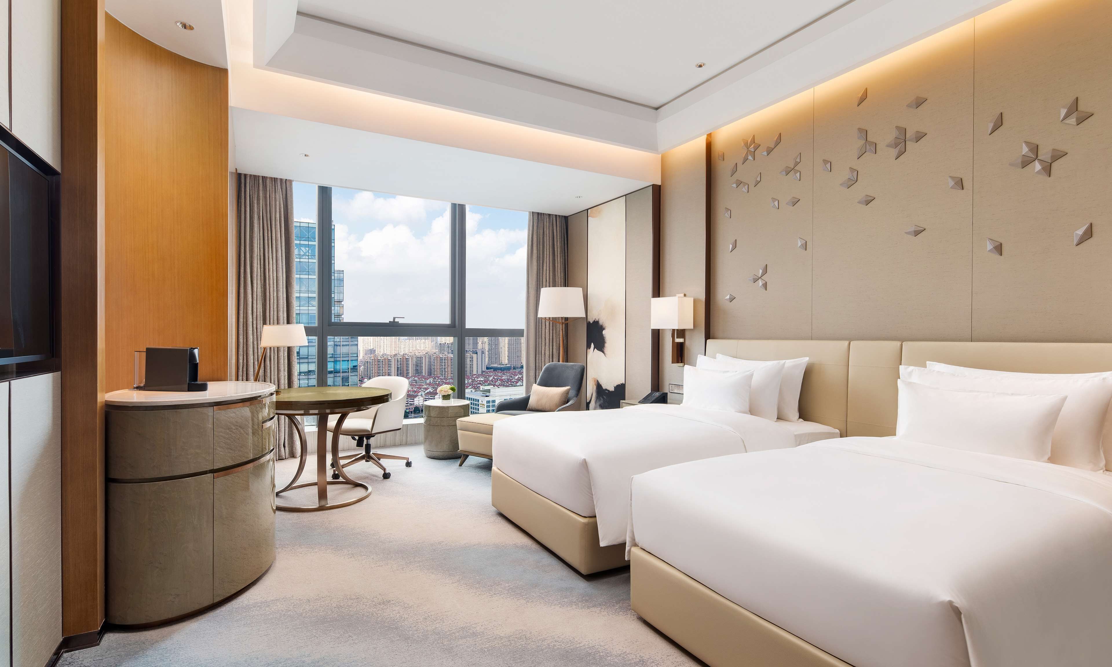 Ein Hotelzimmer mit zwei Betten, einem Schreibtisch, einem Stuhl, einer Lampe und Blick auf die Stadt.