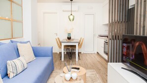Ein modernes Wohnzimmer mit einem blauen Sofa, einem runden Couchtisch mit Dekoration, einem Essbereich mit einem weißen Tisch und Stühlen sowie einem Fernseher auf einem weißen Schrank.