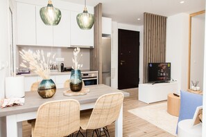 Una cocina moderna con armarios blancos, un comedor con sillas de mimbre y un televisor sobre un soporte blanco.