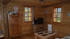 Interior de una cabaña de madera con televisión, muebles de madera y ventanas amplias.