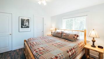 Quarto com colcha xadrez, cabeceira de madeira, mesa de cabeceira com luminária, quadro na parede e janela com vista para árvores.