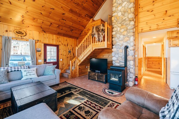 A cozy cabin interior with a wooden staircase, a stone fireplace, a sofa, and a television. - Sunday River Chalet w/ Hot Tub, Fire Pit & River Access Near Ski Lifts (Bethel)