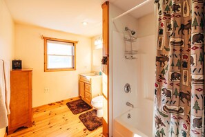 A bathroom with wooden flooring, a window, a toilet, a sink, and a shower with a patterned curtain. - Sunday River Chalet w/ Hot Tub, Fire Pit & River Access Near Ski Lifts (Bethel)