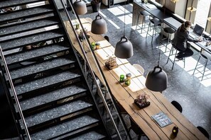 A modern interior with a staircase, long wooden tables, hanging pendant lights, and a person working at a table. - Peak 12 Hotel (Viborg)