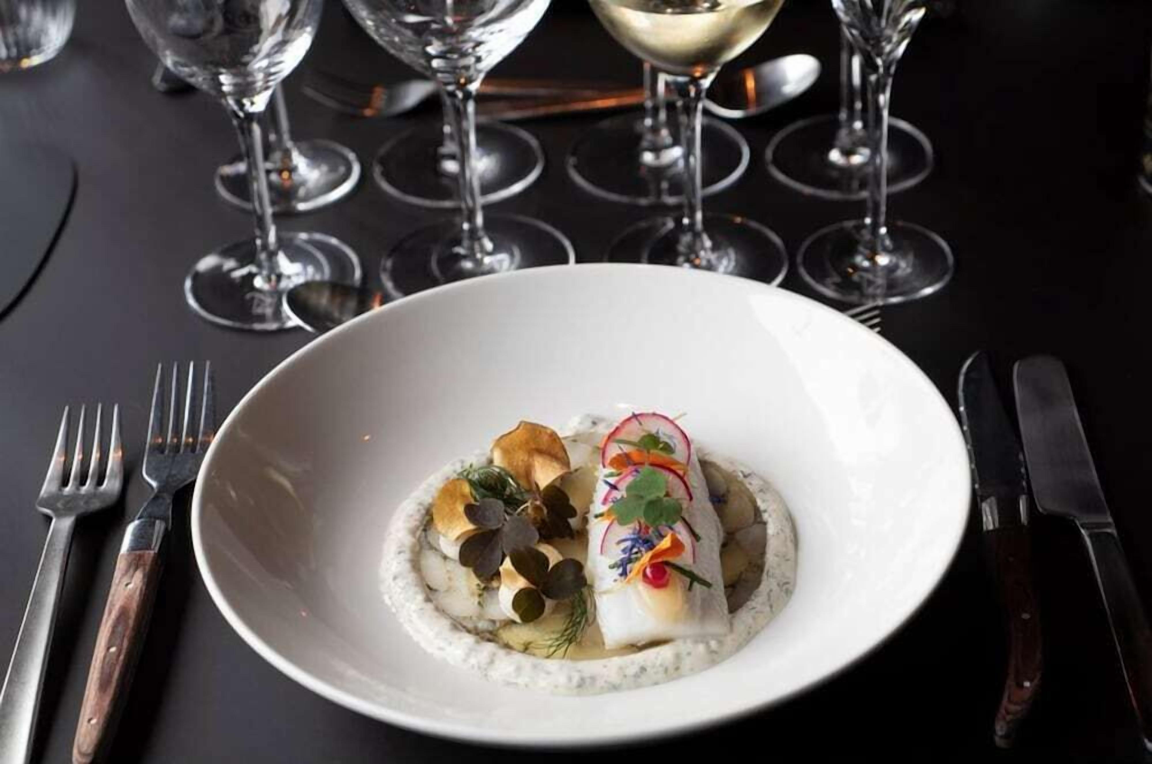 A white plate with a fish dish, garnished with herbs and edible flowers, on a dark table with wine glasses and cutlery.