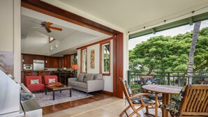A spacious living area with a kitchen, a dining set, and a balcony with a view of trees. - Hale Inu Waina: Spacious 2BR condo in Waikoloa with scenic views. (Waikoaloa)