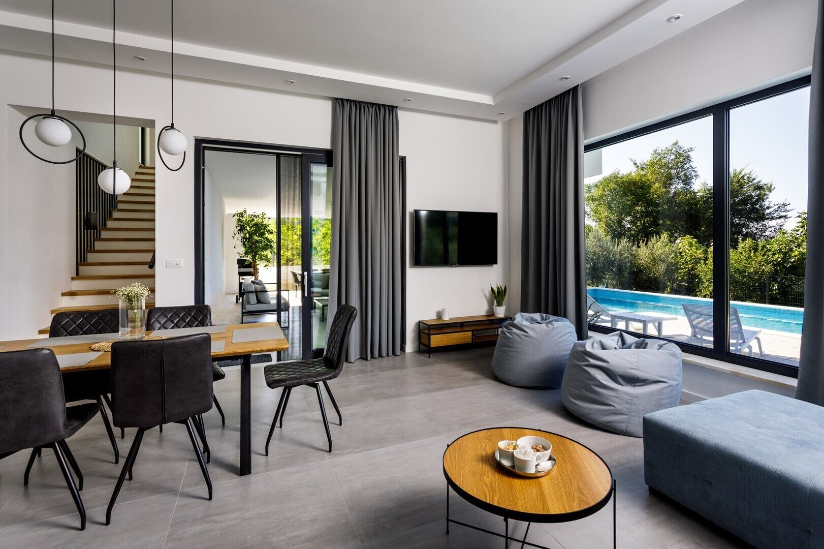 A modern living room with a dining area, a TV mounted on the wall, and a view of a swimming pool.