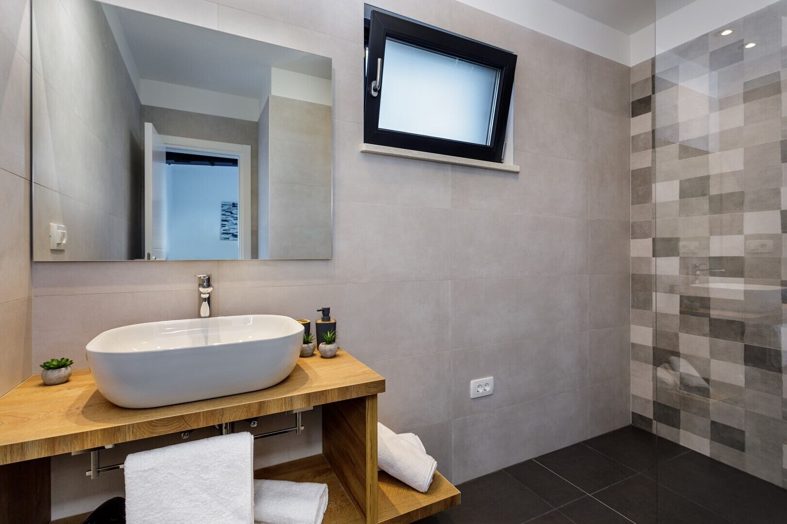 A modern bathroom with a large oval sink, a mirror, a television mounted on the wall, and a tiled shower area.