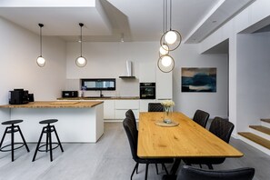 Cozinha moderna com ilha central, banquetas, mesa de jantar com cadeiras, iluminação pendente e um quadro na parede.