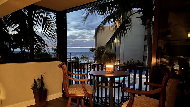 Un balcon avec des chaises et une table en bois, donnant sur une piscine et des palmiers, et offrant une vue sur l’océan.