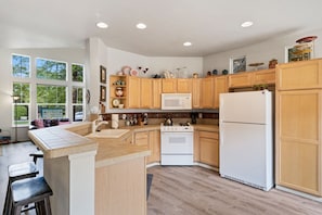 A modern kitchen with wooden cabinets, a white refrigerator, a stove, and a large window overlooking greenery.