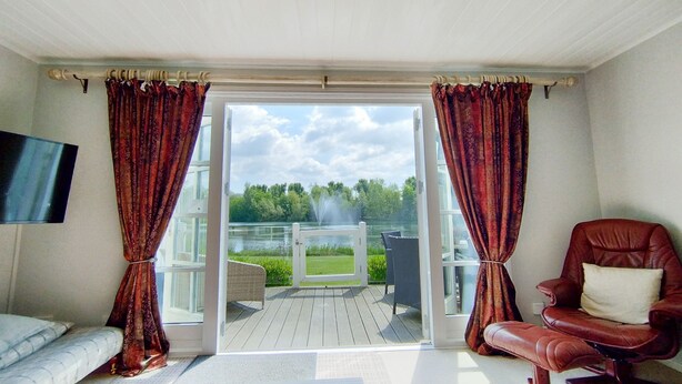 A room with a sliding glass door leading to a deck with a view of a lake and trees. There is a brown leather chair and a white sofa. - Mayfly Lodge - sleeps 6 guests  in 3 bedrooms (Cotswold Lakes)