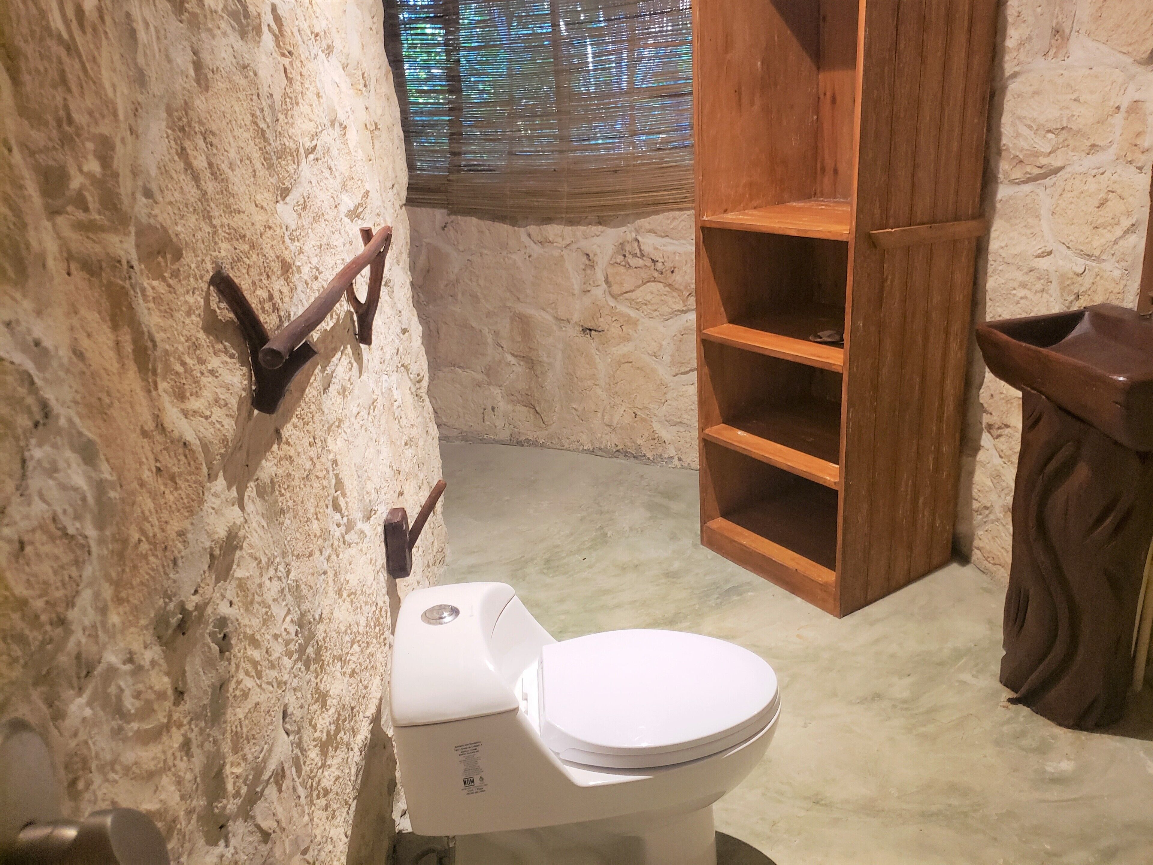 Un baño con inodoro blanco, estantes de madera y paredes de piedra.