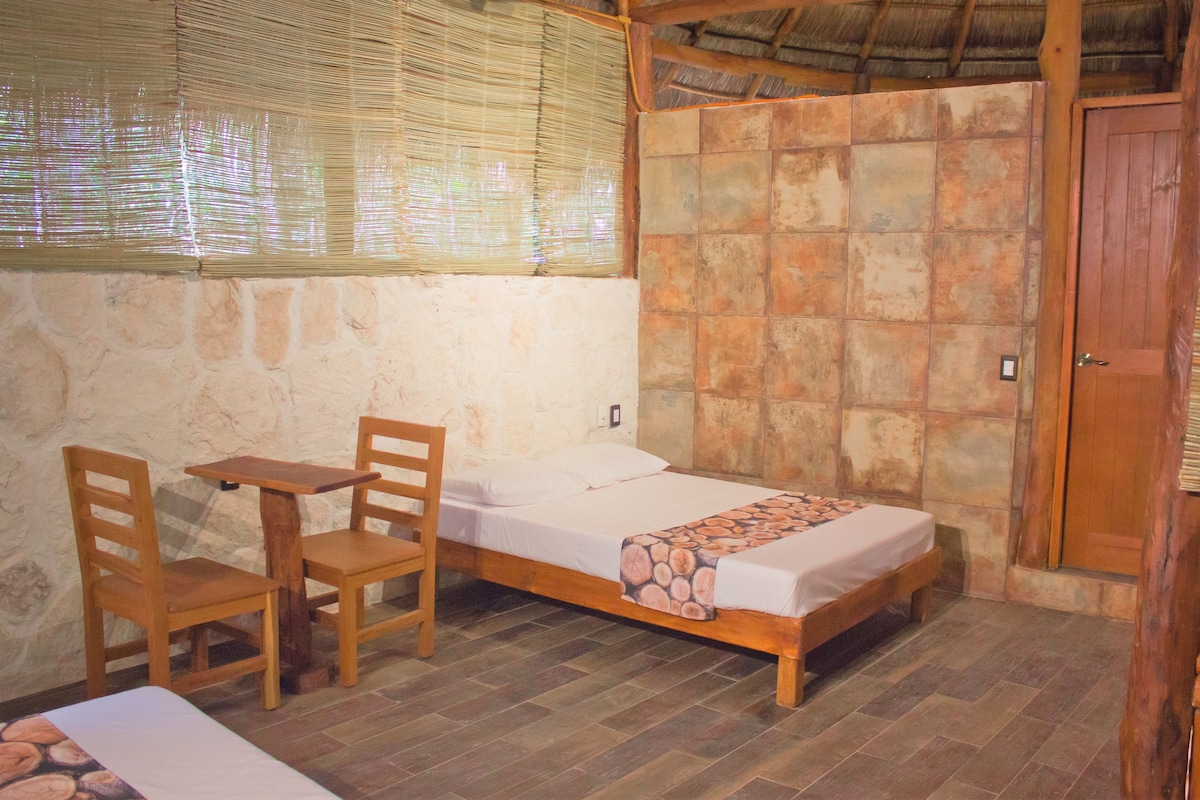 A single bed with a wooden frame and a patterned bedspread, a wooden chair, and a small table in a room with stone walls and a tiled floor.