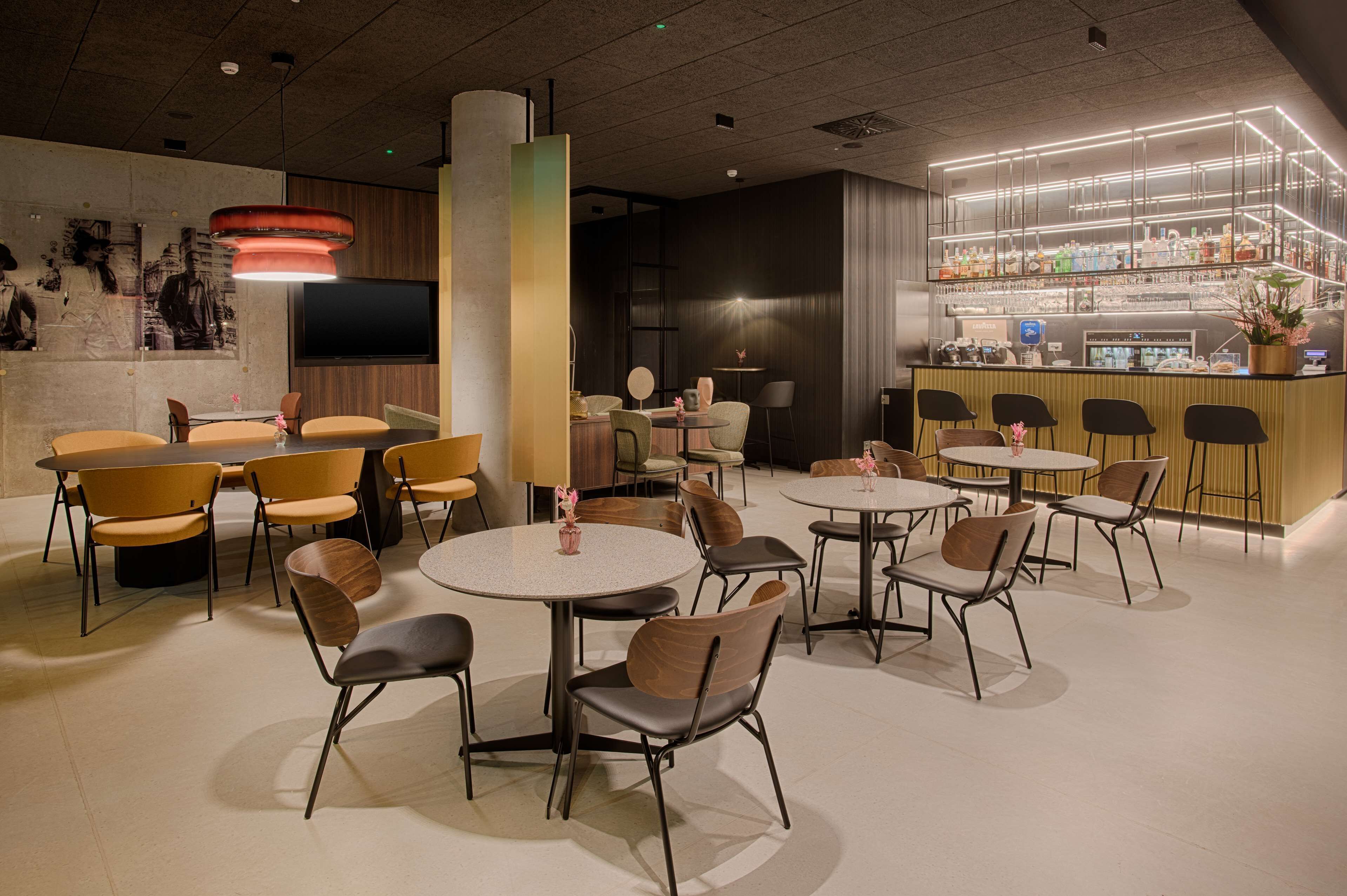 A modern bar area with round tables and chairs, a large TV screen, and a bar counter with various bottles.