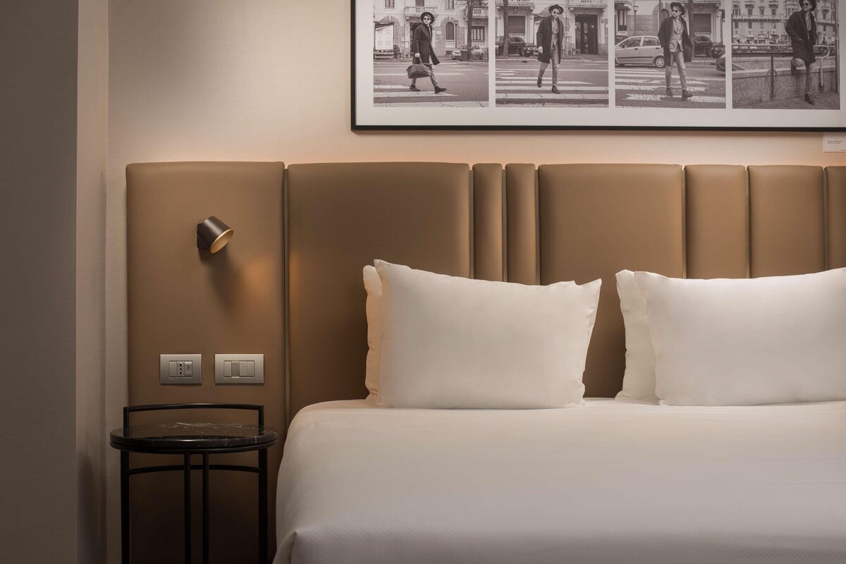 A hotel room with a large bed, a headboard with a light fixture, a small table, and a framed picture on the wall.