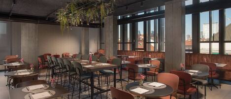 A modern restaurant with round tables and chairs, large windows, and hanging plants.