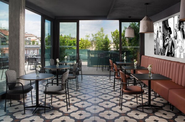 A modern cafe with round tables, cushioned seating, and large windows offering an outdoor view. - NH Milano Corso Buenos Aires  (Milan)