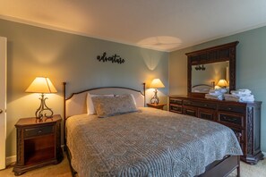 Un lit bien fait, recouvert d’une couvre-lit à motifs, deux lampes de chevet, une commode avec un miroir et un panneau mural sur lequel on peut lire le mot « aventure ».