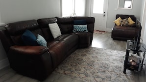 Olohuoneessa on ruskea nahkainen kulmasohva, kuviollinen matto ja televisiotaso, jonka päällä on litteänäyttöinen televisio.