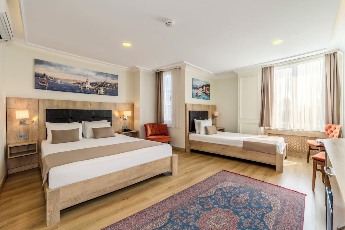 A hotel room with two beds, a desk, a chair, a red armchair, and a large window with curtains.