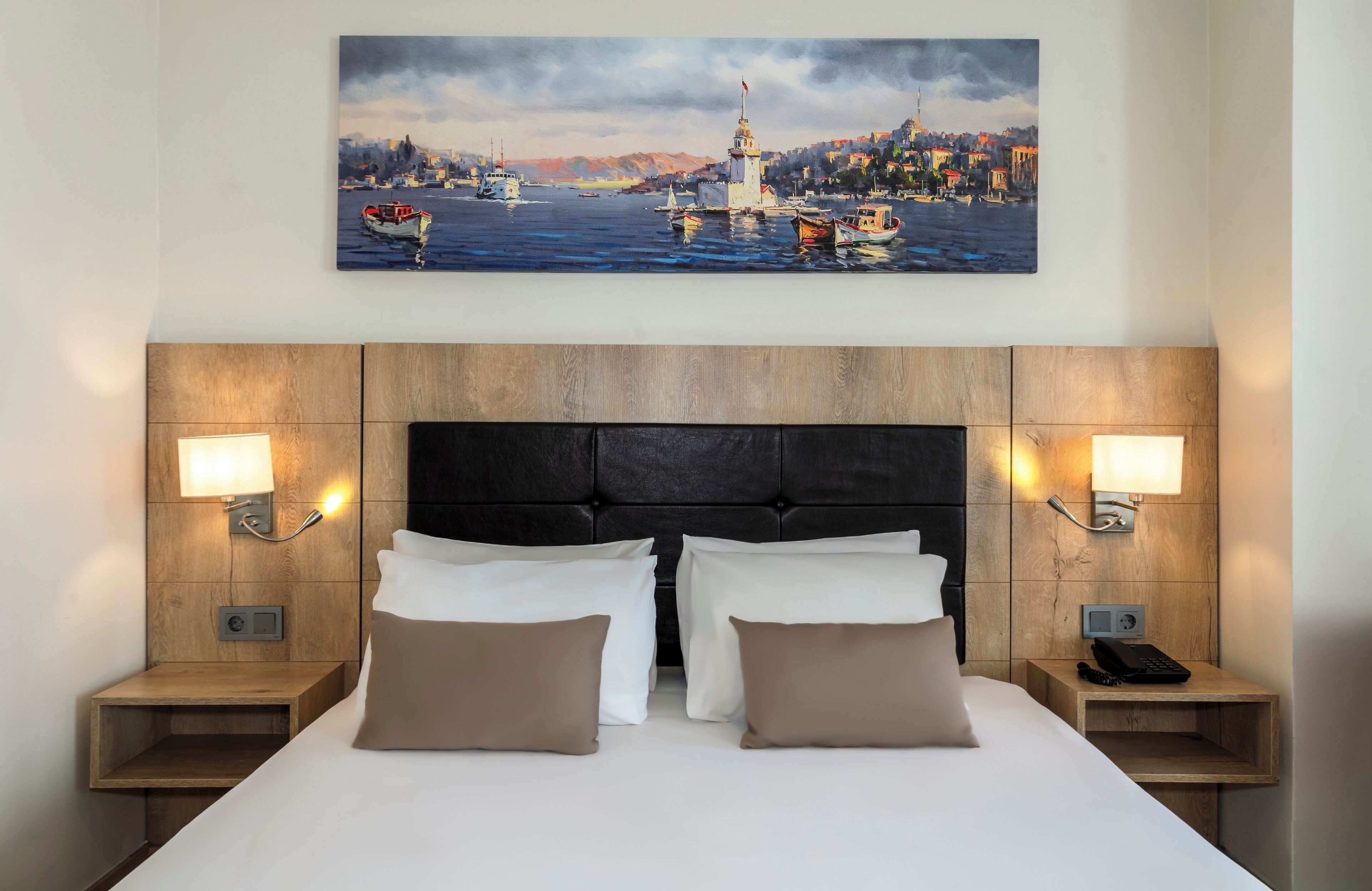 A hotel room with a large bed, two bedside tables, a headboard, and a painting of a coastal scene above the bed.