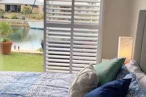 Una camera da letto con un letto, cuscini, una finestra con persiane, vista su un corpo d'acqua e una pianta in vaso.