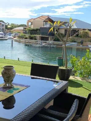A patio with a table, chairs, a potted plant, and a view of a body of water and houses in the background.