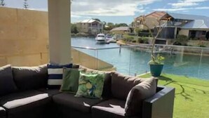 Uma área externa coberta com sofá, mesa de centro, planta em vaso e vista para a água e casas ao fundo.