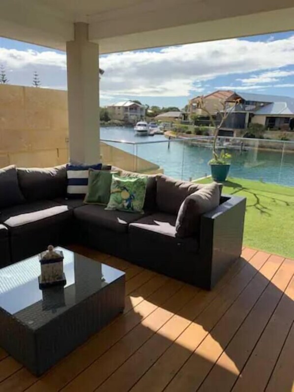 A covered outdoor area with a sofa and coffee table, a potted plant, and a view of a body of water and houses in the background.